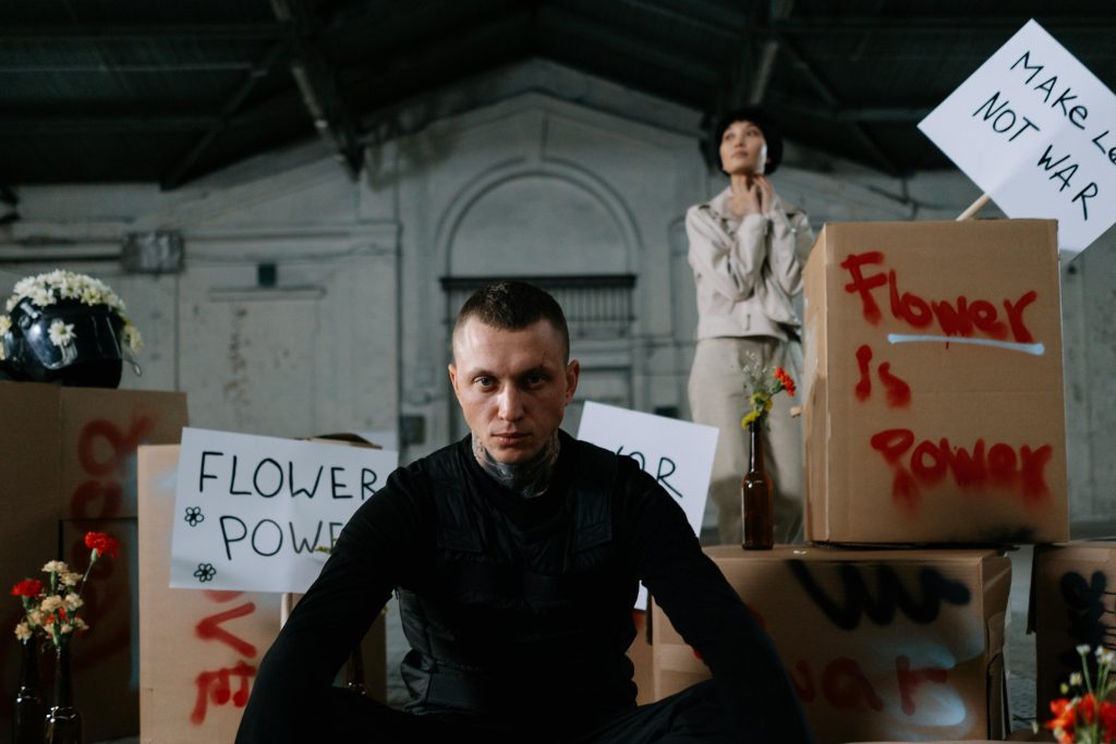 Un homme en noir, assis au premier plan, fixe l'objectif avec intensité. Derrière lui, des pancartes pacifistes et des fleurs évoquent le contraste entre violence intérieure et messages de paix.