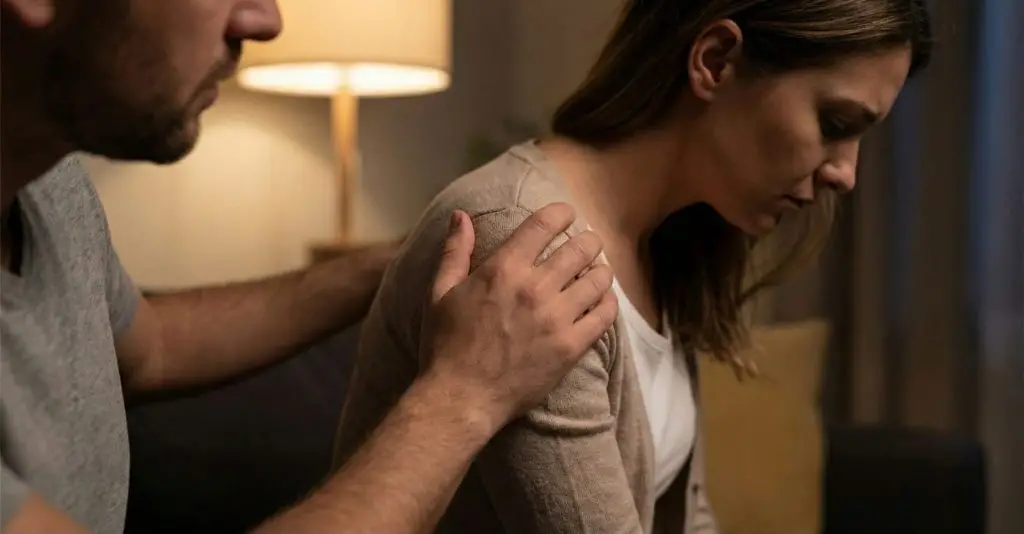 Gros plan sur la main d'un homme posée sur l'épaule tendue d'une femme qui regarde vers le bas, illustrant la saturation sensorielle et le rejet du contact physique dans le couple épuisé.
