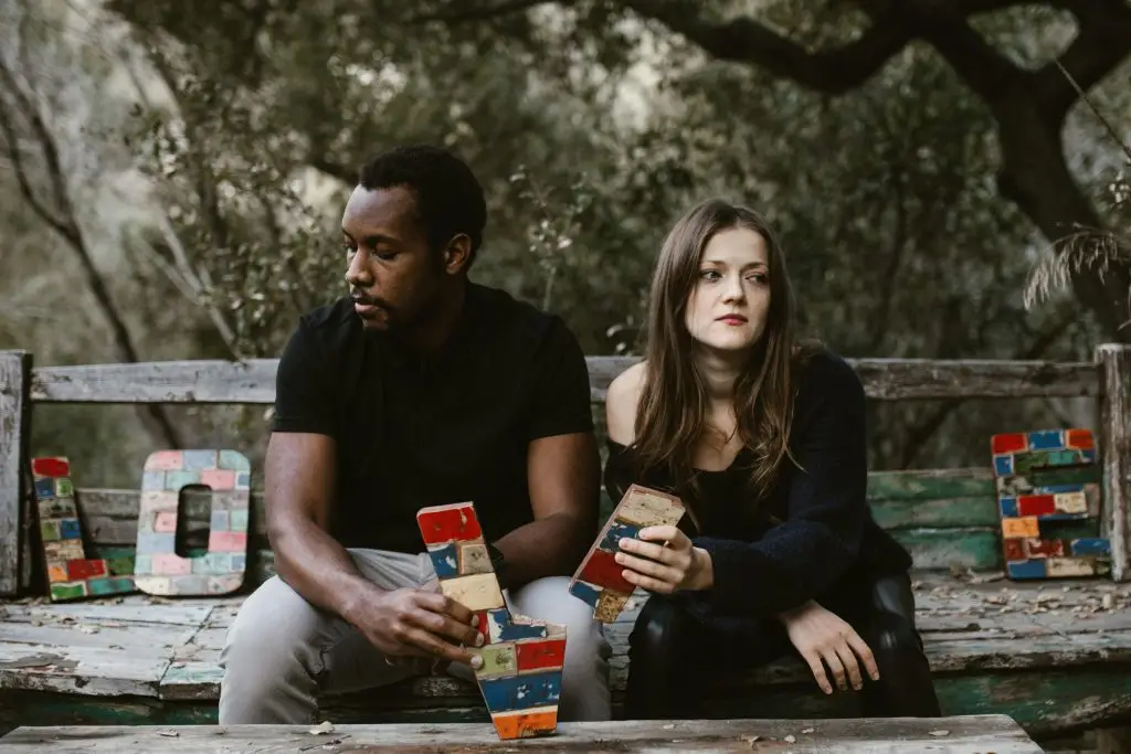 Couple assis sur un banc, détournés l’un de l’autre, tenant une lettre brisée du mot LOVE, illustrant un couple épuisé par le quotidien, la fatigue et des disputes de couple à répétition, sans être un mauvais couple.